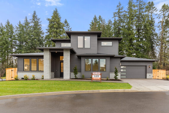 Exterior of 2-story home, dark gray paint with light gray brick accents and columns. Landscaped yard with grass, shrubs, and river rock. For sale sign, built by Glavin Homes