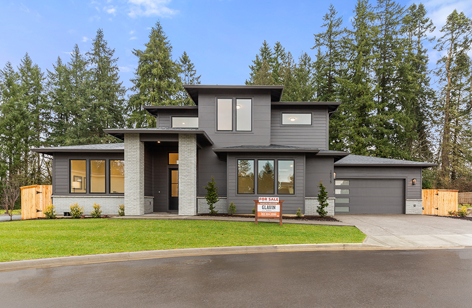 Exterior of 2-story home, dark gray paint with light gray brick accents and columns. Landscaped yard with grass, shrubs, and river rock. For sale sign, built by Glavin Homes